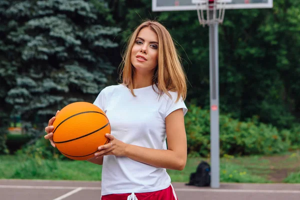 Menina bonita vestida com t-shirt branca, shorts e tênis, joga com uma bola em um campo de basquete . — Fotografia de Stock