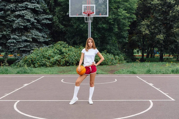 Gadis muda yang cantik mengenakan kaos putih, celana pendek dan sepatu olahraga, bermain dengan bola di lapangan basket . — Stok Foto