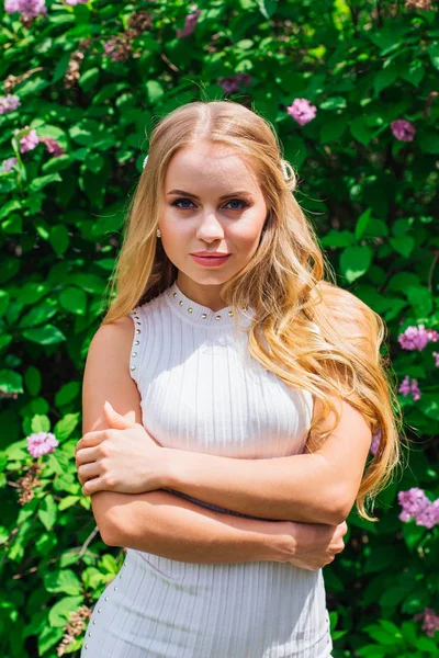 Retrato de una encantadora mujer rubia con hermoso vestido blanco de pie junto al arbusto lila . — Foto de Stock