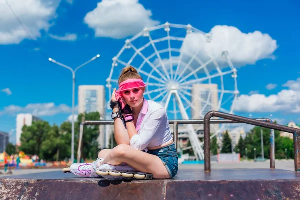 Retrato de uma menina emocional em uma viseira de boné rosa usando luvas de proteção e patins sentados no fundo da roda gigante . — Fotografia de Stock