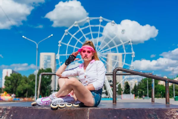 Porträtt av en känslomässig flicka i en rosa mössa visor bär skyddshandskar och rullskridskor sitter på bakgrunden av pariserhjulet. — Stockfoto