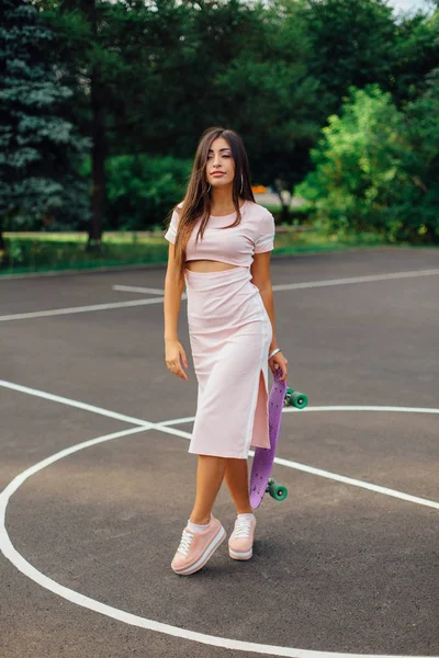 Portret van een lachende charmante brunette vrouw houdt haar skateboard op een basketbalveld. — Stockfoto