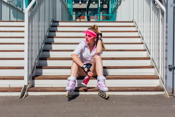 Porträtt av en känslomässig flicka i en rosa mössa visor bär skyddshandskar och rullskridskor sitter på trappor hålla en flaska vatten i händerna — Stockfoto