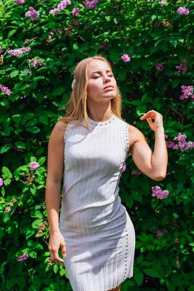 Porträt einer charmanten blonden Frau in schönem weißen Kleid, die neben Fliederbüschen steht. — Stockfoto