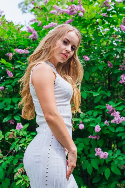 Retrato de uma mulher loira encantadora vestindo belo vestido branco em pé ao lado de arbusto lilás . — Fotografia de Stock