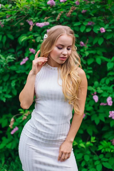 Retrato de una encantadora mujer rubia con hermoso vestido blanco de pie junto al arbusto lila . — Foto de Stock