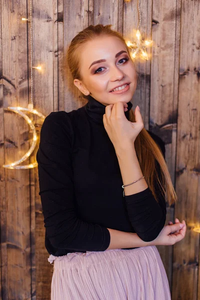 Όμορφη νεαρή γυναίκα ντυμένη σε ένα πουλόβερ και φούστα στέκεται δίπλα στο ξύλινο τοίχο με αστέρια και φεγγίτες φώτα — Φωτογραφία Αρχείου