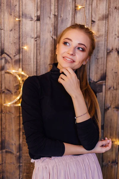 Όμορφη νεαρή γυναίκα ντυμένη σε ένα πουλόβερ και φούστα στέκεται δίπλα στο ξύλινο τοίχο με αστέρια και φεγγίτες φώτα — Φωτογραφία Αρχείου