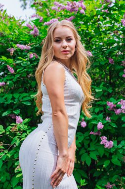 Leylak çalıyanında duran güzel beyaz elbise giyen büyüleyici sarışın bir kadın portresi.