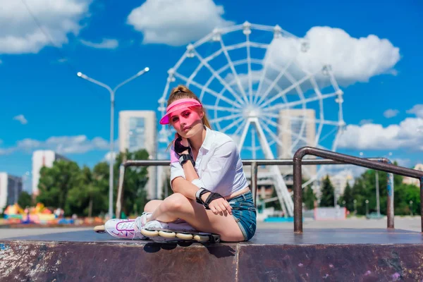 Retrato de uma menina emocional em uma viseira de boné rosa usando luvas de proteção e patins sentados no fundo da roda gigante . — Fotografia de Stock