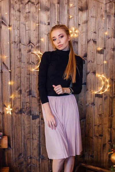 Hermosa joven vestida con un suéter y falda de pie junto a la pared de madera con luces de estrella y luna —  Fotos de Stock