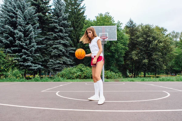 Piękna młoda dziewczyna ubrana w biały t-shirt, szorty i trampki, gra w piłkę na boisko do koszykówki. — Zdjęcie stockowe