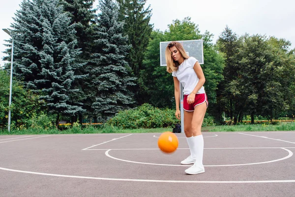 Hermosa joven vestida con camiseta blanca, pantalones cortos y zapatillas de deporte, juega con una pelota en una cancha de baloncesto . —  Fotos de Stock