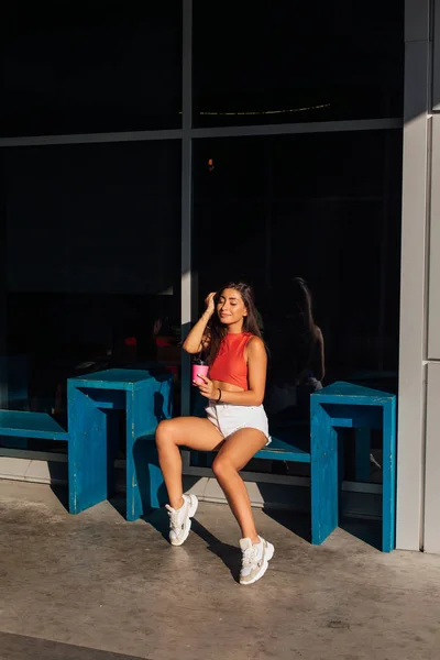 Elegante mujer morena feliz joven con pantalones cortos blancos y zapatillas de deporte con taza de café rosa para llevar . — Foto de Stock