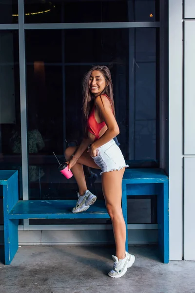 Elegante mujer morena feliz joven con pantalones cortos blancos y zapatillas de deporte con taza de café rosa para llevar . — Foto de Stock