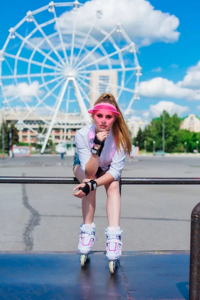 Portret van een emotioneel meisje in een roze GLB Visor dragen beschermende handschoenen en inline skates zittend op de achtergrond van reuzenrad. — Stockfoto