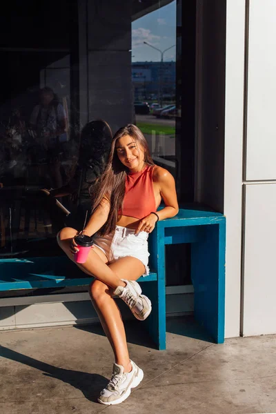 Elegante feliz jovem morena mulher vestindo branco shorts e tênis segurando rosa xícara de café para ir . Imagem De Stock