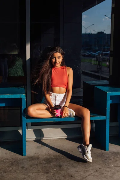 Elegante mujer morena feliz joven con pantalones cortos blancos y zapatillas de deporte con taza de café rosa para llevar . —  Fotos de Stock