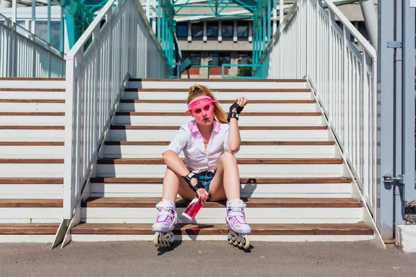 Potret seorang gadis emosional dengan topi pink mengenakan sarung tangan pelindung dan rollerblades duduk di tangga sambil memegang botol air di tangan — Stok Foto