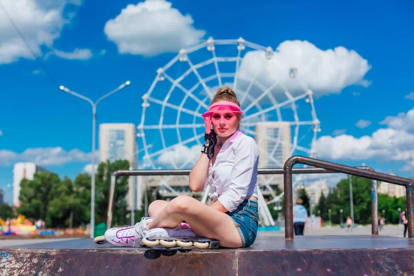 Porträtt av en känslomässig flicka i en rosa mössa visor bär skyddshandskar och rullskridskor sitter på bakgrunden av pariserhjulet. — Stockfoto