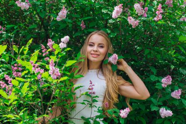 Leylak çalıyanında duran güzel beyaz elbise giyen büyüleyici sarışın bir kadın portresi.