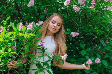 Leylak çalıyanında duran güzel beyaz elbise giyen büyüleyici sarışın bir kadın portresi.