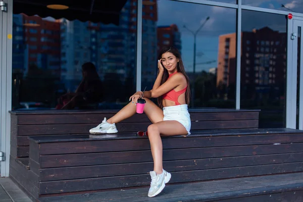 Elegante feliz jovem morena mulher vestindo branco shorts e tênis segurando rosa xícara de café para ir sentado ao lado de café . — Fotografia de Stock