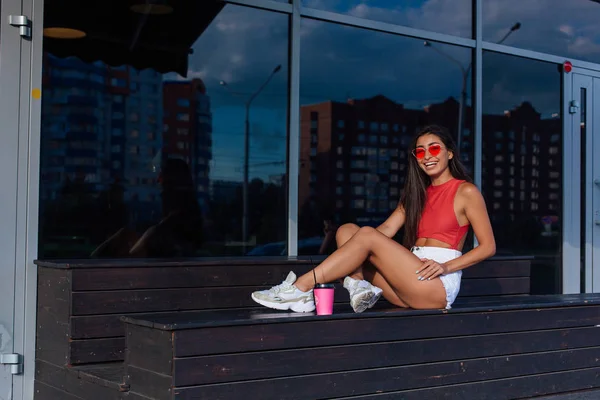 Elegante feliz jovem morena mulher vestindo branco shorts e tênis segurando rosa xícara de café para ir sentado ao lado de café . — Fotografia de Stock