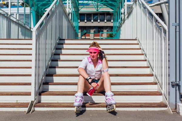 Potret seorang gadis emosional dengan topi pink mengenakan sarung tangan pelindung dan rollerblades duduk di tangga sambil memegang botol air di tangan — Stok Foto