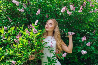 Leylak çalıyanında duran güzel beyaz elbise giyen büyüleyici sarışın bir kadın portresi.