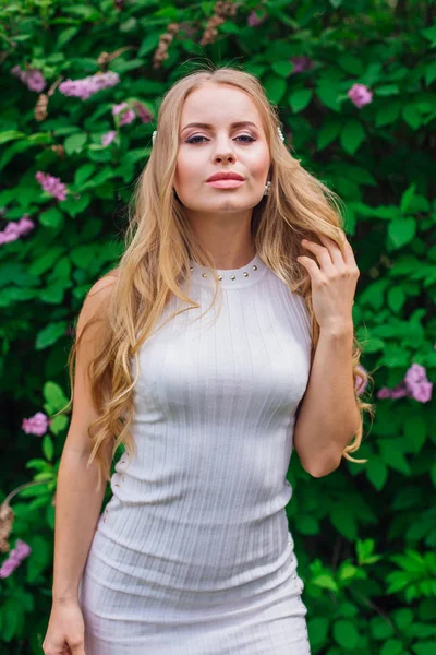 Портрет чарівної блондинки в красивій білій сукні, що стоїть поруч з бузковим кущем . — стокове фото