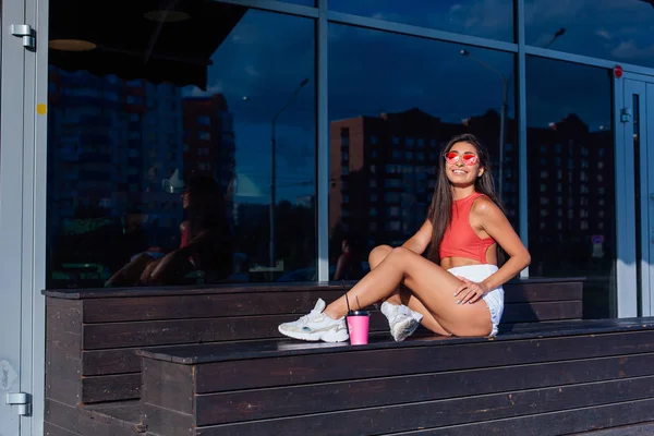 Elegante feliz jovem morena mulher vestindo branco shorts e tênis segurando rosa xícara de café para ir sentado ao lado de café . — Fotografia de Stock