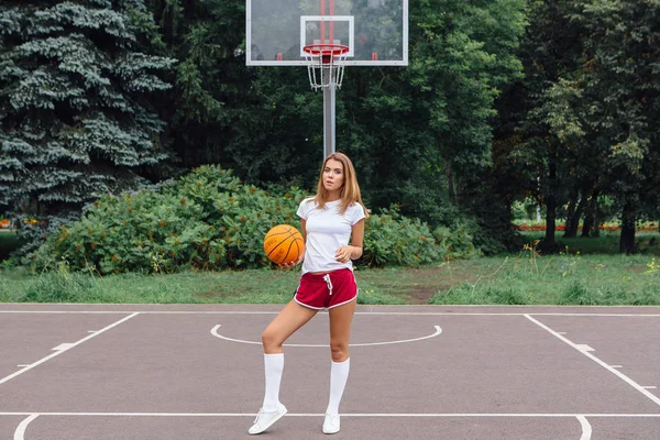白の t シャツ、ショート パンツ、スニーカー、服を着た美しい少女はバスケット ボール コートでボールで遊ぶ. — ストック写真