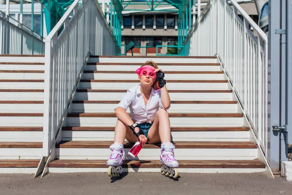 Retrato de uma menina emocional em um visor de boné rosa usando luvas de proteção e patins sentados em escadas segurando uma garrafa de água nas mãos — Fotografia de Stock