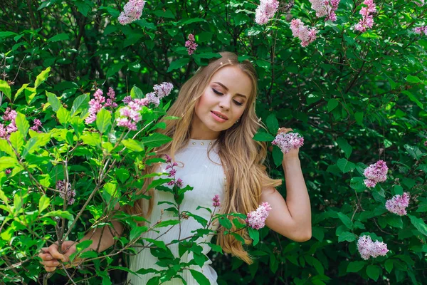 Leylak çalıyanında duran güzel beyaz elbise giyen büyüleyici sarışın bir kadın portresi.