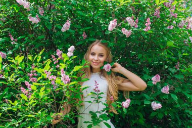 Leylak çalıyanında duran güzel beyaz elbise giyen büyüleyici sarışın bir kadın portresi.