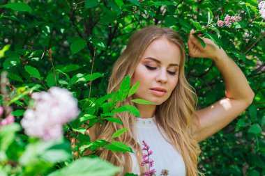 Leylak çalıyanında duran güzel beyaz elbise giyen büyüleyici sarışın bir kadın portresi.