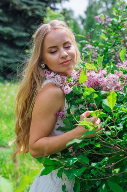 Leylak çalıyanında duran güzel beyaz elbise giyen büyüleyici sarışın bir kadın portresi.