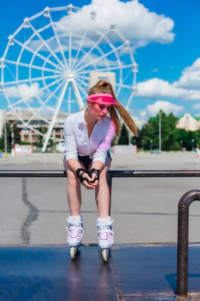Портрет емоційної дівчини в рожевому ковпачку в захисних рукавичках і роликових лопатях, що сидять на фоні коліщатка . — стокове фото