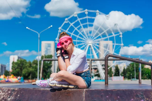 Potret seorang gadis emosional dengan topi merah muda mengenakan sarung tangan pelindung dan rollerblades duduk di latar belakang roda ferris . — Stok Foto