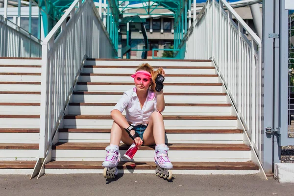 Porträtt av en känslomässig flicka i en rosa mössa visor bär skyddshandskar och rullskridskor sitter på trappor hålla en flaska vatten i händerna — Stockfoto
