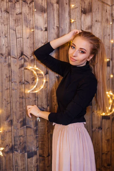 Hermosa joven vestida con un suéter y falda de pie junto a la pared de madera con luces de estrella y luna — Foto de Stock