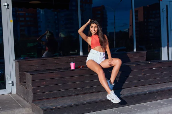 Snygg lycklig ung brunett kvinna klädd i vita shorts och sneakers håller rosa kopp kaffe att gå bredvid kafé. — Stockfoto