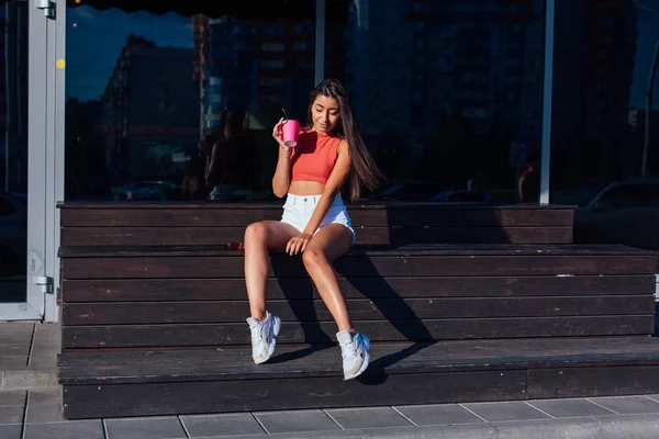 Elegante feliz jovem morena mulher vestindo branco shorts e tênis segurando rosa xícara de café para ir sentado ao lado de café . — Fotografia de Stock