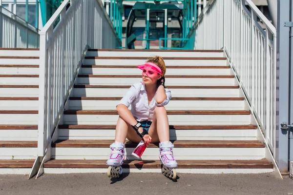 Portrait d'une fille émotionnelle dans une visière à capuchon rose portant des gants de protection et des patins à roulettes assis sur des escaliers tenant une bouteille d'eau dans les mains — Photo