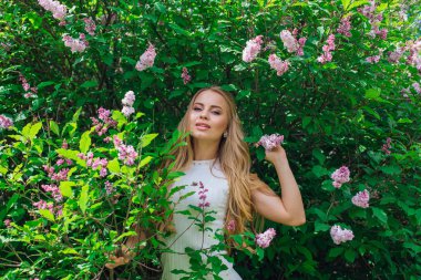 Leylak çalıyanında duran güzel beyaz elbise giyen büyüleyici sarışın bir kadın portresi.