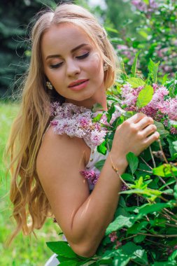 Leylak çalıyanında duran güzel beyaz elbise giyen büyüleyici sarışın bir kadın portresi.