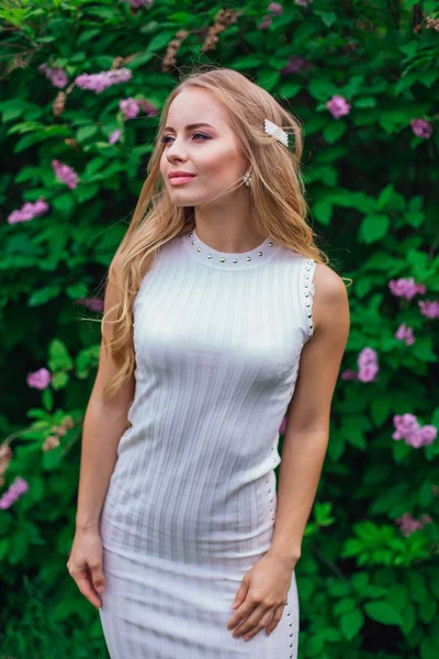 Retrato de una encantadora mujer rubia con hermoso vestido blanco de pie junto al arbusto lila . — Foto de Stock