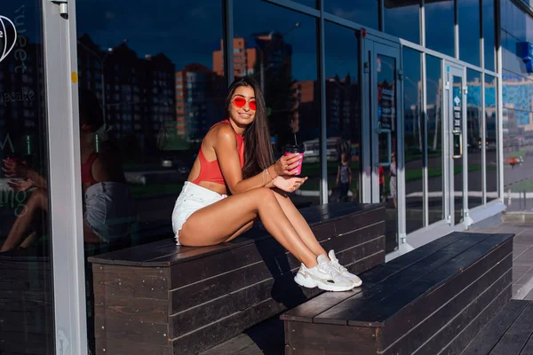 Elegante feliz jovem morena mulher vestindo branco shorts e tênis segurando rosa xícara de café para ir sentado ao lado de café . — Fotografia de Stock