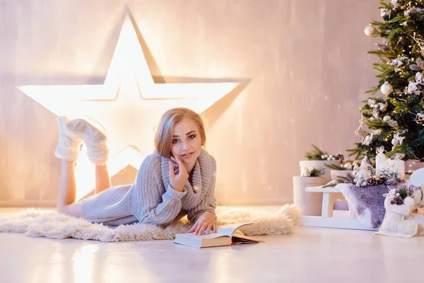 Beautiful young woman laying and reading book under Christmas tree in a holiday interior — Stock Photo, Image